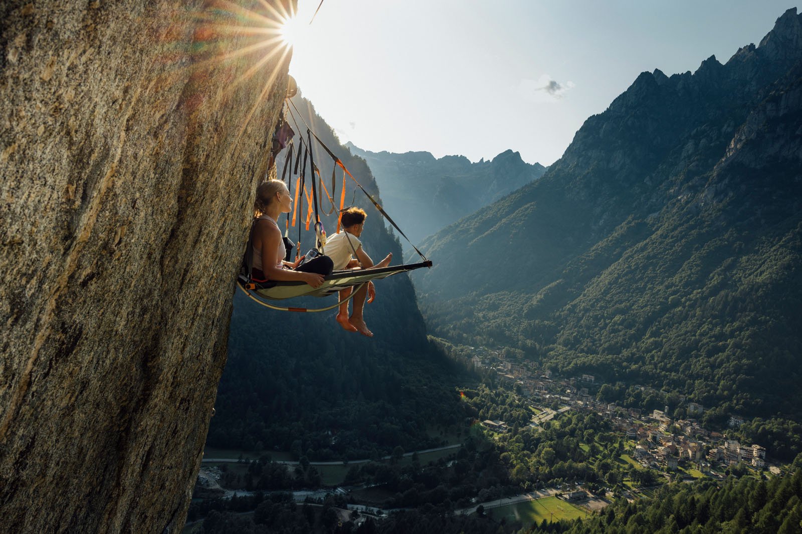 Two people sit on a hanging platform attached to a steep cliff, overlooking a lush valley and distant mountains under a bright sun. The scene is adventurous and peaceful, with a village visible far below.