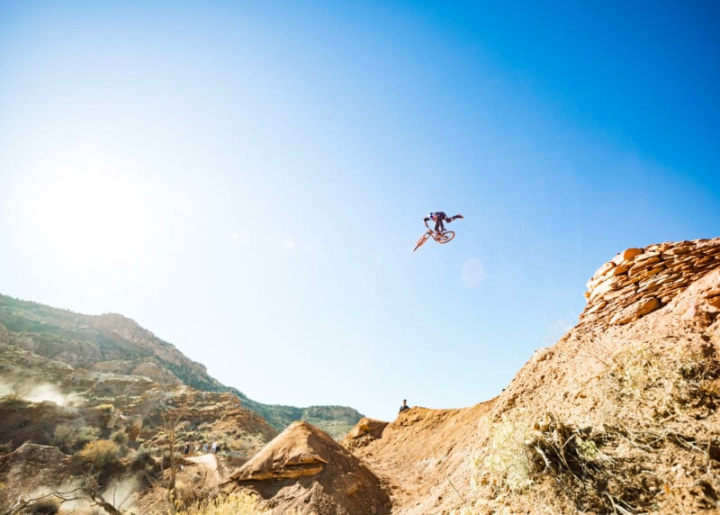 A mountain biker performs a high jump off a dirt ramp in a rocky, desert landscape under a clear blue sky, with sunlight shining from the left.