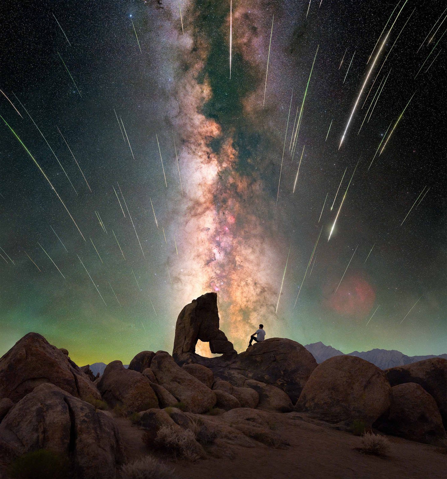 A person sits on rocky terrain beneath a stone arch, gazing at the vibrant Milky Way and numerous shooting stars streaking across the night sky.