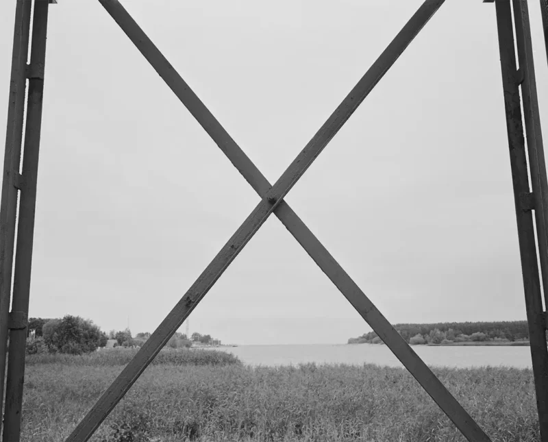 A large metal X-shaped structure frames a view of a river or lake bordered by grass and trees under an overcast sky.