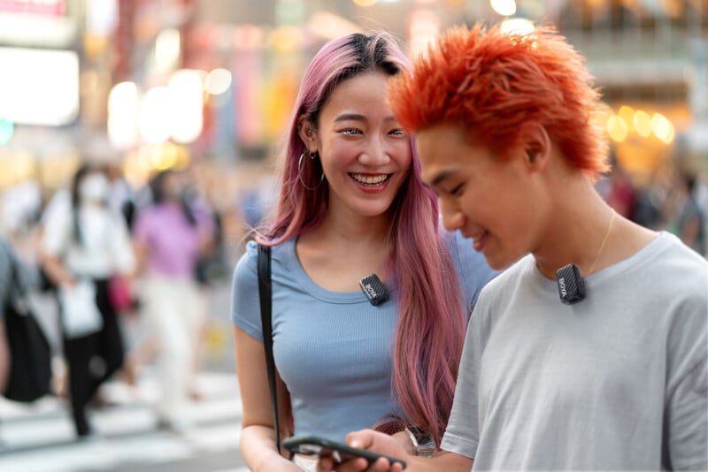 Two young people with colorful hair and clip-on microphones stand in a lively city street. The woman with long pink hair smiles while the man with short orange hair looks down at his phone. The background is blurred with crowds.