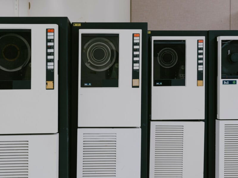 Three vintage mainframe computer units with circular disk drives and control panels, white front panels, and black frames are lined up side by side against a neutral background.