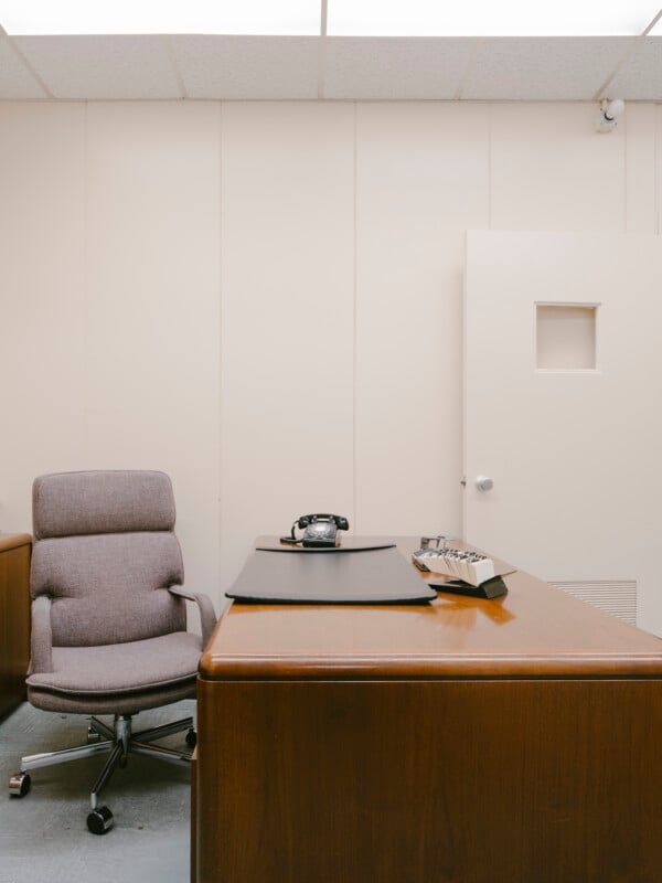 A minimalist office with a wooden desk, a gray upholstered chair, a rotary phone, and a desk organizer. The walls are plain and light-colored, and a door with a small square window is partially open in the background.