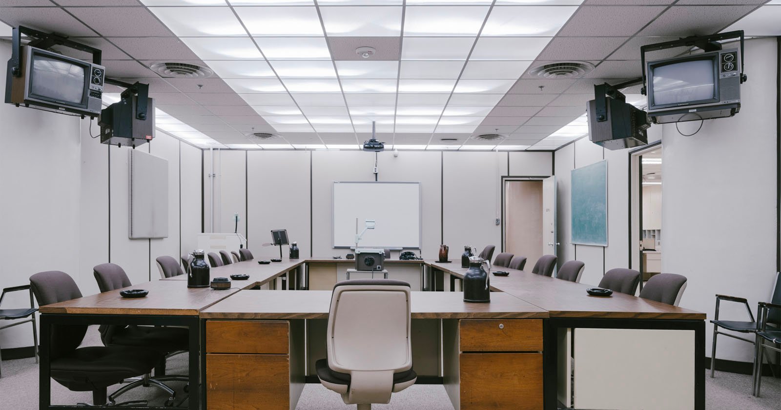 A large, empty conference room with a square arrangement of desks and brown office chairs, four ceiling-mounted CRT televisions, a whiteboard, and fluorescent lighting overhead.