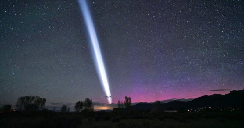 A bright white spotlight beams into a star-filled night sky, with purple and pink auroras glowing above silhouetted trees and distant mountains.
