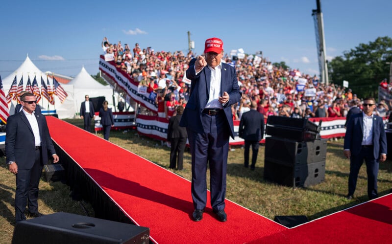 A man in a red "Make America Great Again" hat stands on a red carpet at an outdoor rally, pointing toward the camera, with a large crowd and American flags in the background.