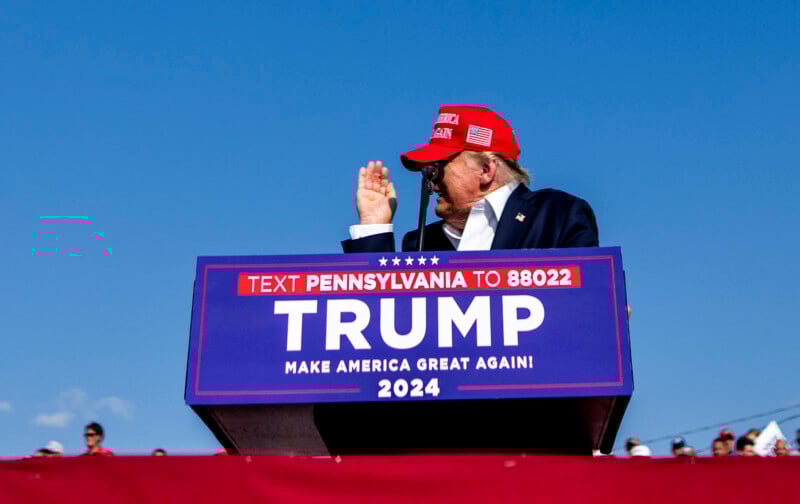 A man in a red "Make America Great Again" hat stands behind a podium reading "Text Pennsylvania to 88022, Trump, Make America Great Again! 2024," speaking at an outdoor event under a clear blue sky.