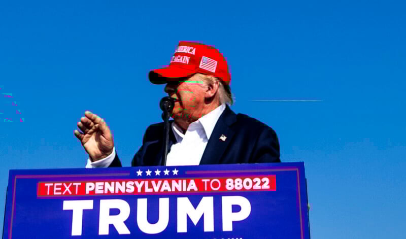 A man in a red "Make America Great Again" hat speaks at a podium with a sign reading "Text PENNSYLVANIA to 88022 TRUMP" against a clear blue sky.