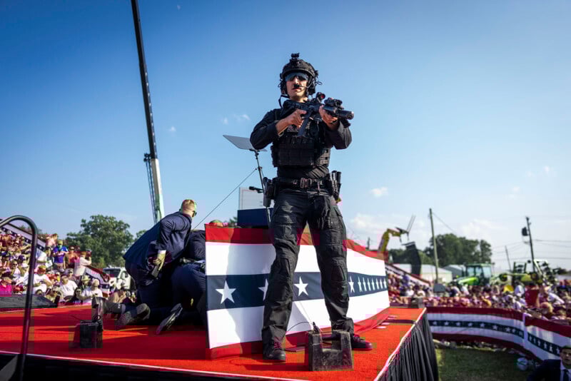 A heavily armed security officer in tactical gear stands alert on a stage decorated with red, white, and blue bunting at an outdoor event, while a man crouches nearby and crowds watch in the background.