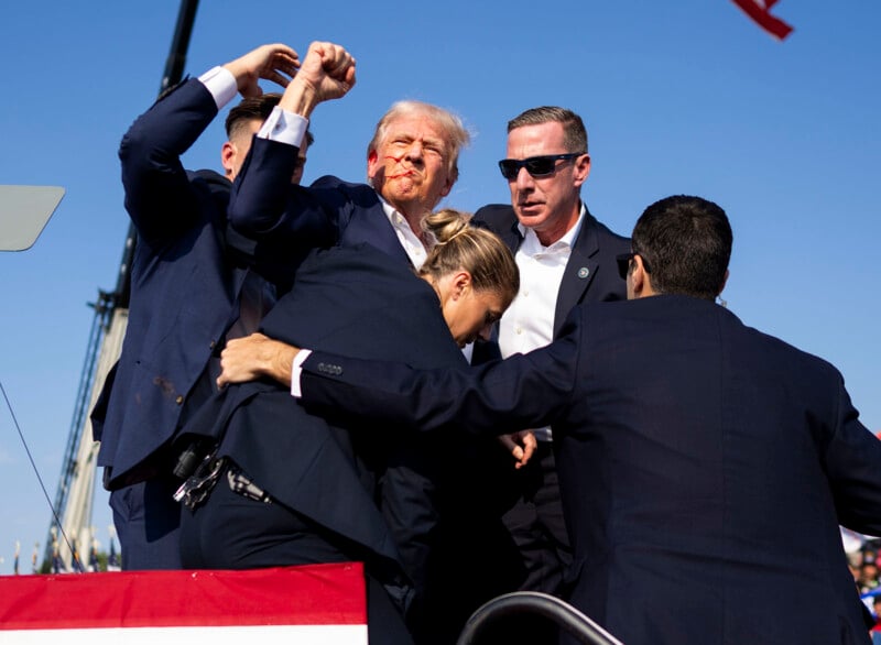 A man with a bloody face is surrounded and shielded by several people in suits, possibly security personnel, at an outdoor event under a clear blue sky.