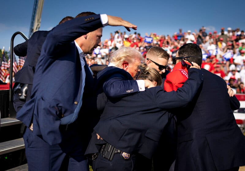 A group of security personnel urgently surround and shield a man with blond hair at an outdoor event, with a large crowd and American flags visible in the background.