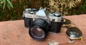 A vintage Canon AE-1 film camera sits on a stone surface next to a pair of aviator sunglasses, with green bamboo plants in the background.