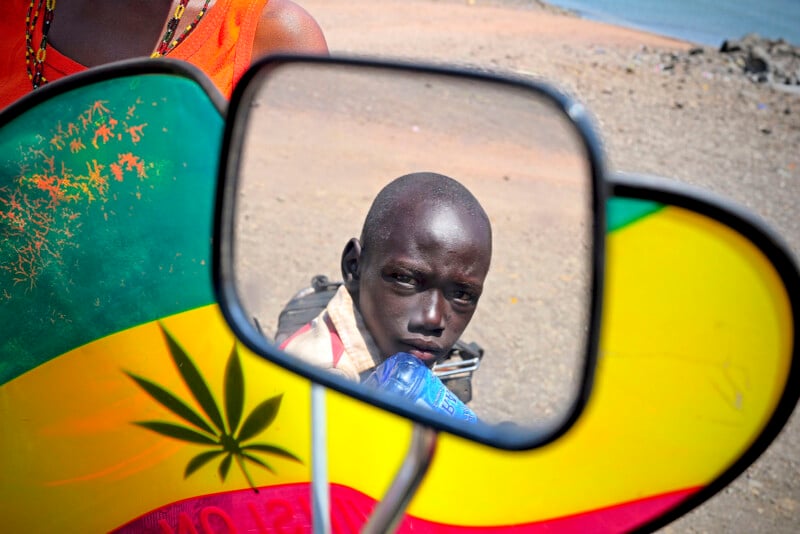 A young boy's face is reflected in a motorcycle mirror, with a colorful panel featuring a large marijuana leaf in the background. The setting appears outdoors on a dirt road near water.