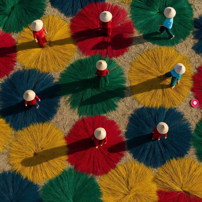 Aerial view of people in conical hats standing among large, colorful bundles of dyed reeds or fibers arranged in circular patterns on the ground. The bundles are red, green, blue, and yellow.