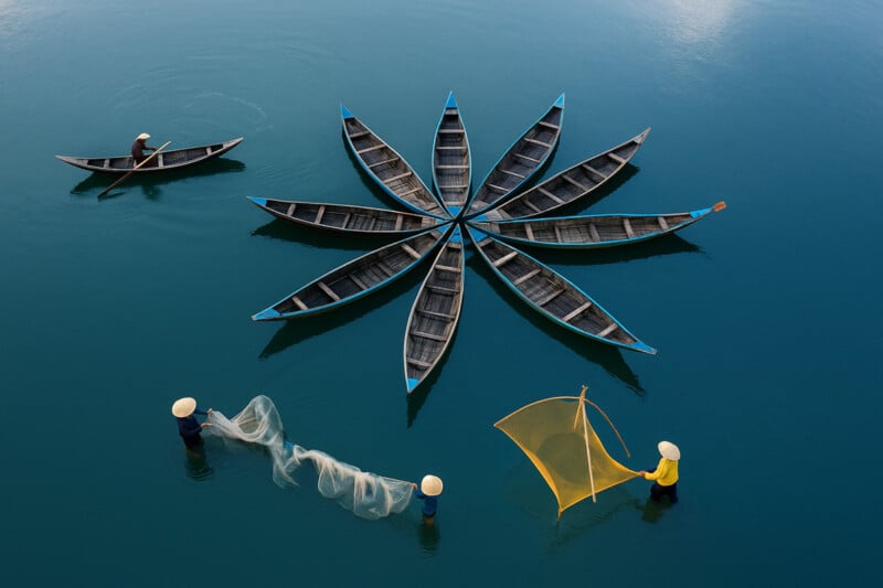 Aerial view of boats arranged in a flower shape on calm blue water, with three people nearby—two wading and casting nets, and one paddling a boat.