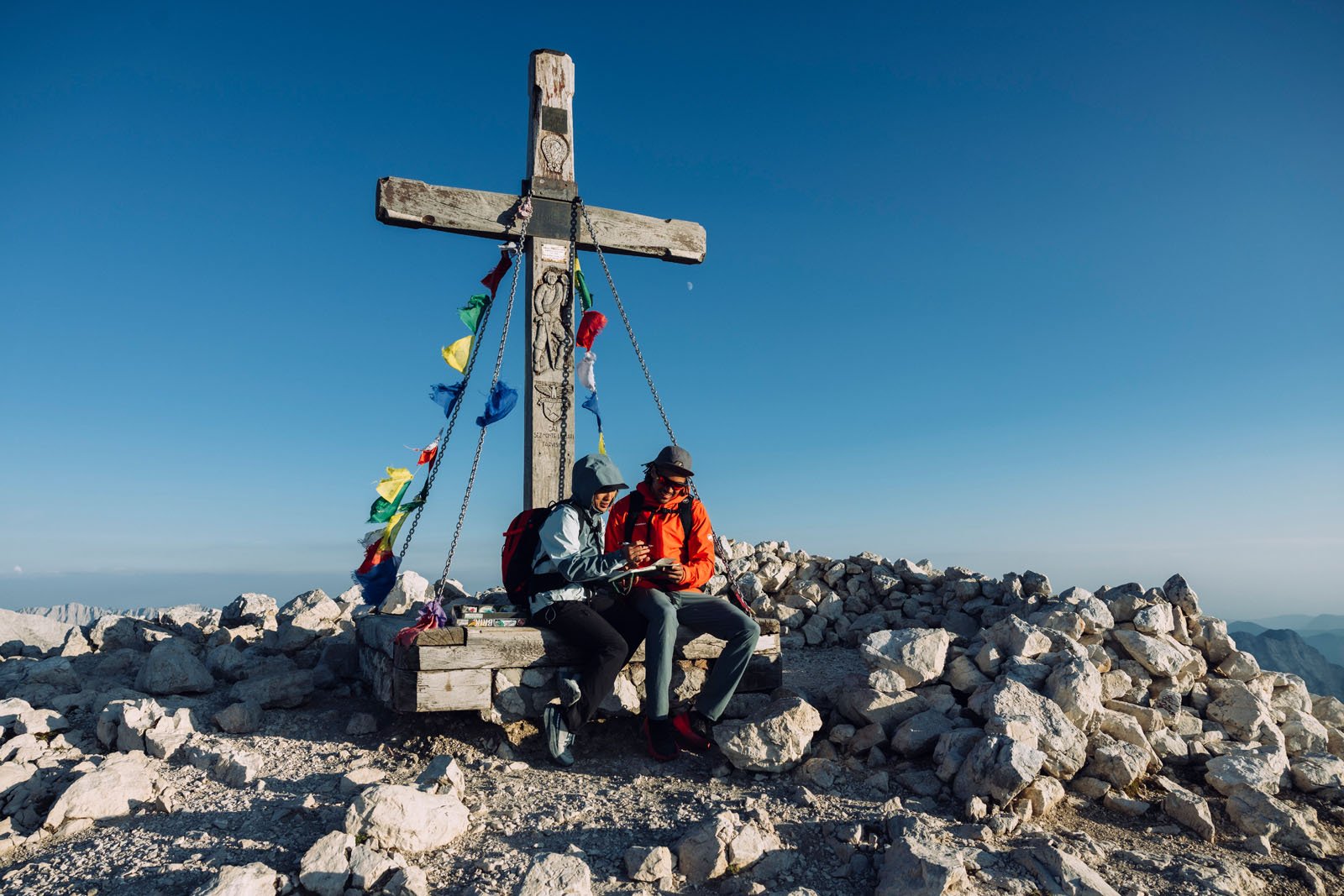 Two hikers sit beside a large wooden cross adorned with prayer flags at a rocky mountain summit under a clear blue sky, looking at something together.