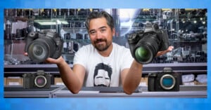 A man with a beard and mustache stands behind a counter in a camera shop, holding a Canon camera in one hand and a Nikon camera in the other. A Fujifilm and a Lumix camera are displayed in front of him.