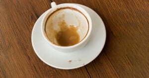 A nearly empty white coffee cup with coffee residue sits on a matching saucer, placed on a wooden table. Some coffee stains are visible on the cup rim and the saucer.