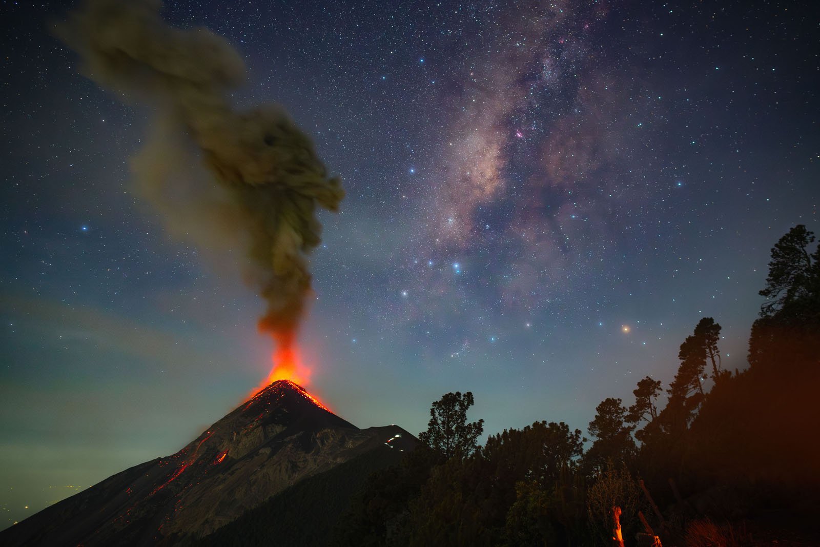 A volcano erupts at night, spewing smoke and lava under a clear, star-filled sky with the Milky Way visible. Silhouetted trees frame the scene in the foreground.