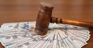 A wooden judge’s gavel rests on top of a spread of one hundred dollar bills, symbolizing the intersection of law and money.
