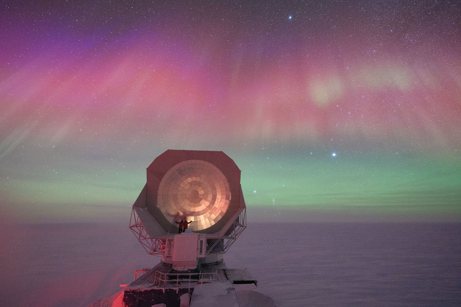 A large radio telescope dish stands on a snowy landscape under a night sky filled with vibrant pink, purple, and green auroras. The area is illuminated by the soft glow of the northern lights.