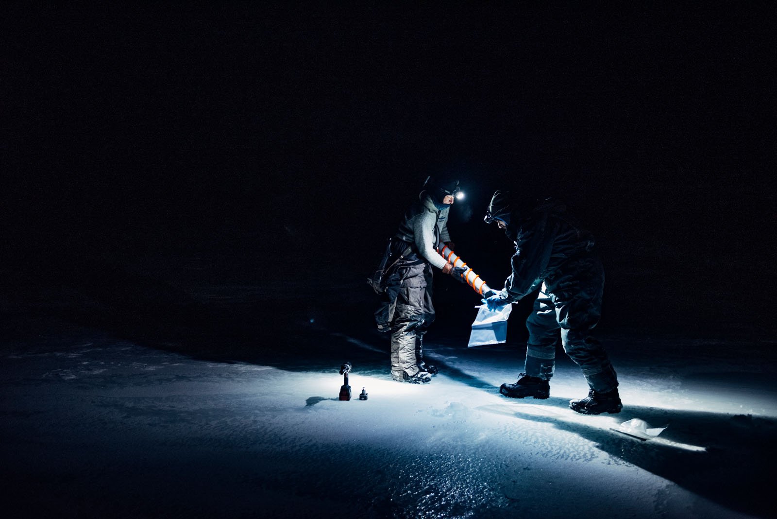 Two people wearing headlamps and winter gear work together on a snowy or icy surface at night, illuminated by their lights in an otherwise dark environment.