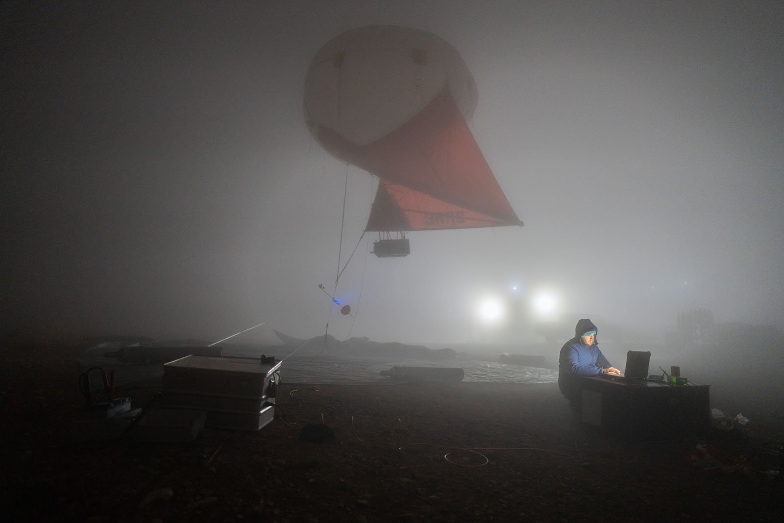 A person in a blue jacket works on a laptop at night in a foggy outdoor setting, illuminated by bright lights, with a large scientific balloon and equipment in the background.