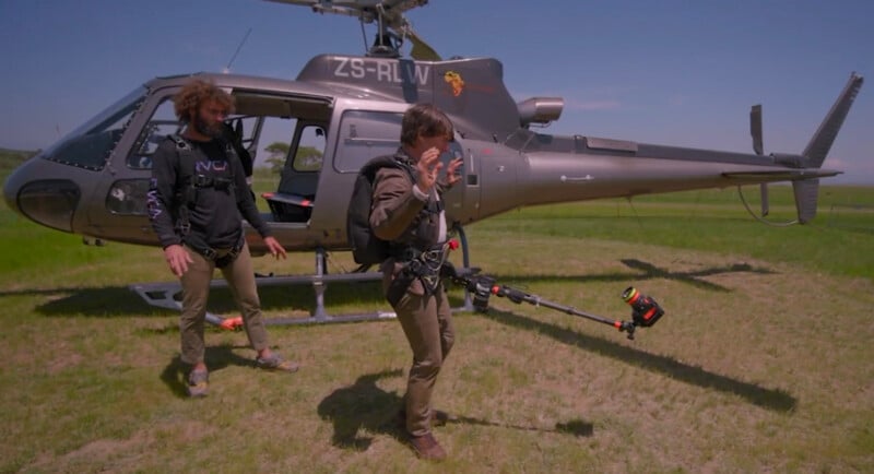 Two people in outdoor gear stand beside a parked helicopter on grass. One person carries a large pole-mounted camera rig; the other stands nearby. It appears to be a film or documentary shoot on a sunny day.