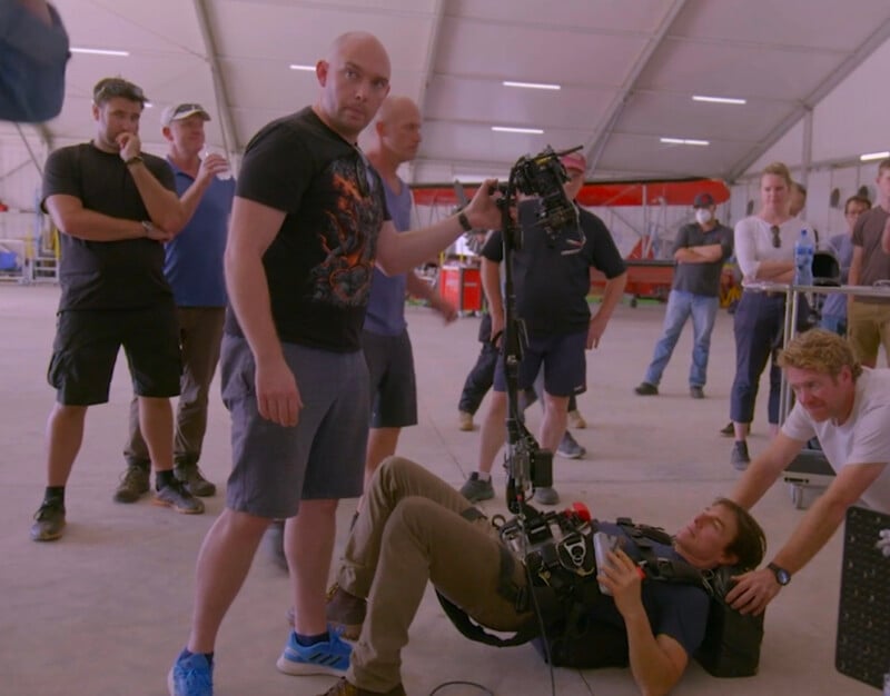 A group of people stand inside a large tent, observing a man lying on the floor operating a camera rig, while another man in shorts and a black T-shirt holds equipment and looks at the camera.
