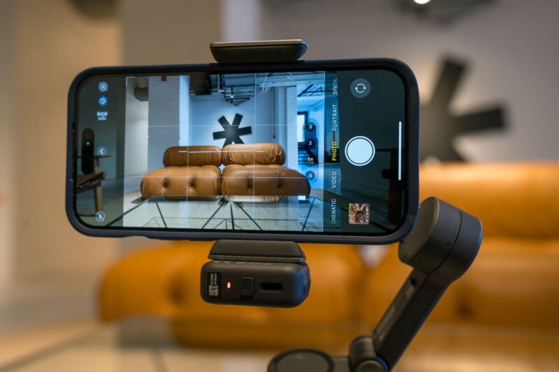 A smartphone mounted on a tripod captures a photo of a modern room with a brown leather couch and large abstract wall art in the background. The phone screen displays the camera interface.