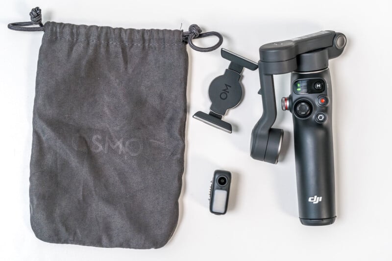 A black DJI handheld gimbal, a smartphone clamp, a small gray and black accessory, and a gray drawstring pouch are arranged on a white surface.