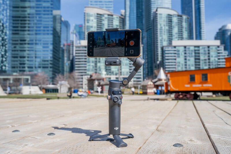 A smartphone mounted on a handheld gimbal tripod records video outdoors, with tall modern buildings and a bright orange train car in the background on a sunny day.