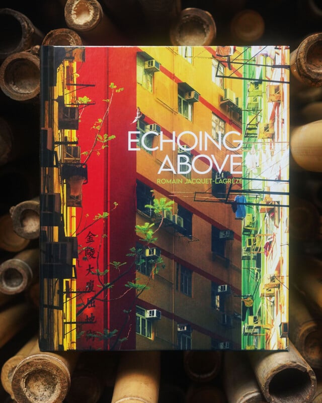 A book titled "Echoing Above" by Romain Jacquet-Lagrèze, featuring a vibrant cover with red and yellow building facades and green balconies, is placed on a background of stacked bamboo poles.