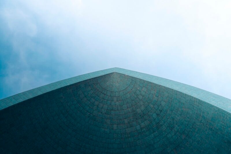A close-up view of the pointed corner of a modern, tiled building against a pale, cloudy sky, photographed from below to highlight its geometric and symmetrical lines.