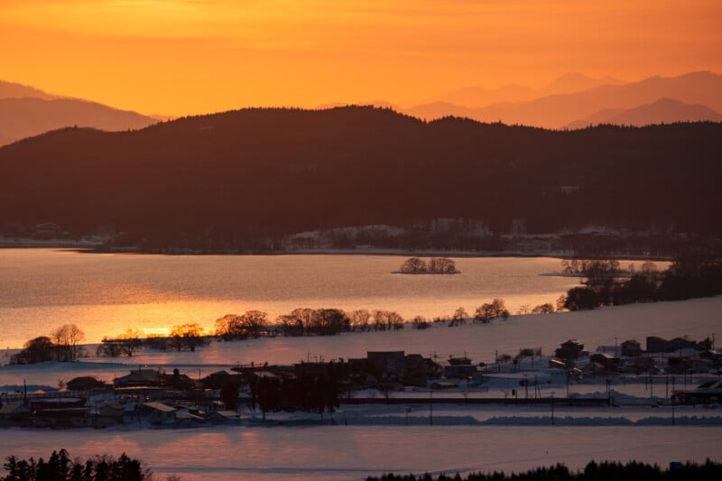 A tranquil sunset over a snowy village by a lake, with silhouettes of trees and mountains in the background, and orange hues reflecting on the water and sky.