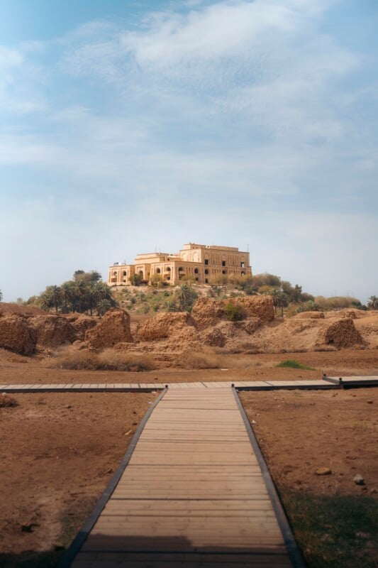 A wooden walkway leads across dry, rocky terrain to a large, historic yellow building atop a hill, surrounded by sparse vegetation under a partly cloudy sky.