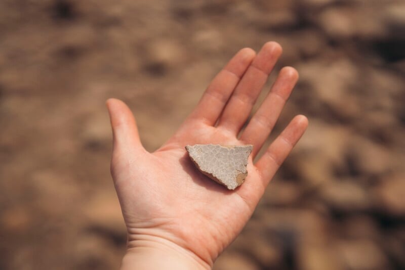 A person’s outstretched hand holds a small, jagged piece of stone outdoors, with a blurred, rocky background.