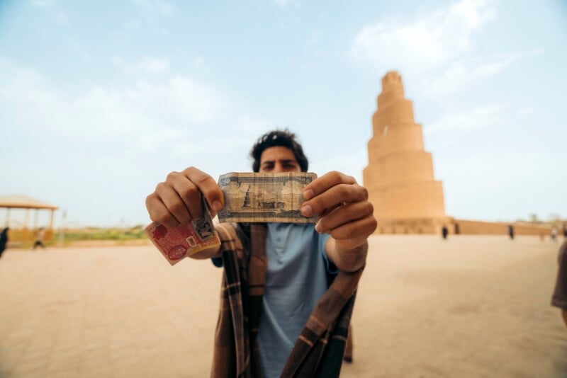 A person standing outdoors holds up a banknote and a card toward the camera, with the Malwiya Tower, a spiral minaret, visible in the background under a partly cloudy sky.
