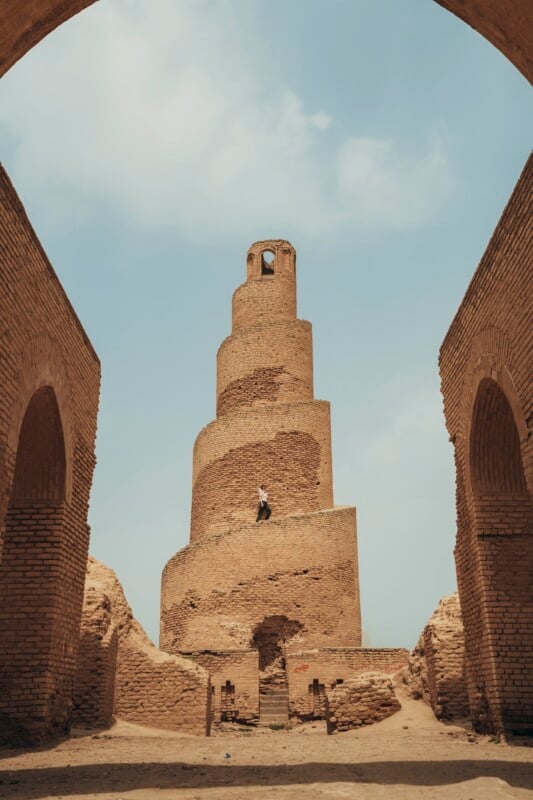 A person stands on the spiral ramp of the ancient brick Malwiya Tower of the Great Mosque of Samarra in Iraq, framed by two large arched brick walls under a partly cloudy blue sky.