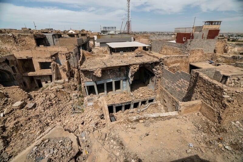 Aerial view of heavily damaged, collapsed buildings in a city. Rubble and debris cover the ground, with walls and roofs partially destroyed. The area looks deserted and affected by conflict or disaster.
