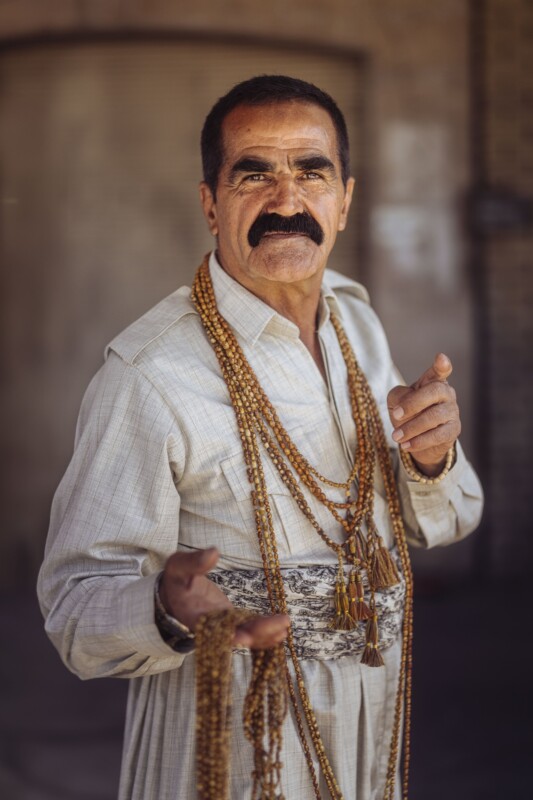 A man with a thick mustache wears a light traditional outfit and multiple long beaded necklaces. He stands outdoors, gesturing with one hand while holding more beads in the other, looking confidently at the camera.