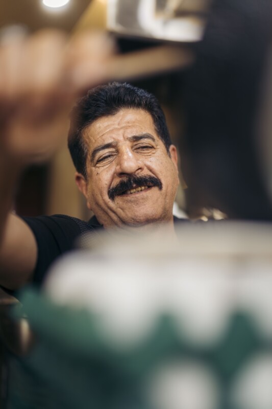 A man with a mustache smiles warmly while reaching out with his arm. The background is softly blurred, drawing focus to his cheerful expression.