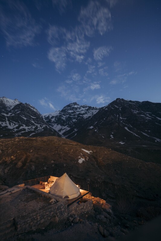 A warmly lit tent sits on a rocky hill at dusk, surrounded by snow-capped mountains under a clear, starry sky. The landscape is serene and remote, with soft evening light highlighting the peaks.