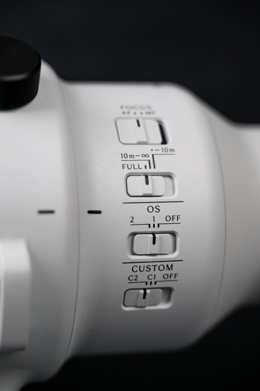 Close-up of a white camera lens with several labeled switches for focus mode, focus range, optical stabilization (OS), and custom settings, set against a dark background.