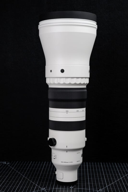 A large white telephoto camera lens with black accents stands upright against a black background, showcasing its adjustable zoom rings and detailed markings.
