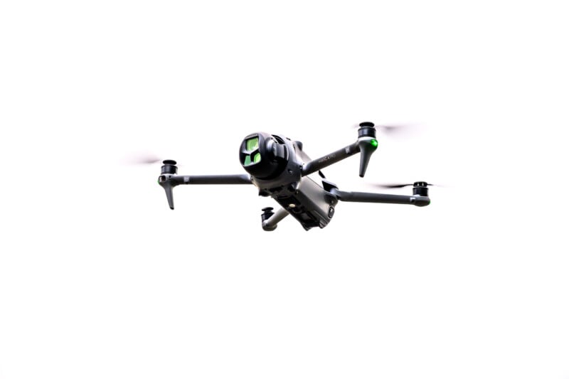 A black quadcopter drone with a camera and green lights is flying against a white background.