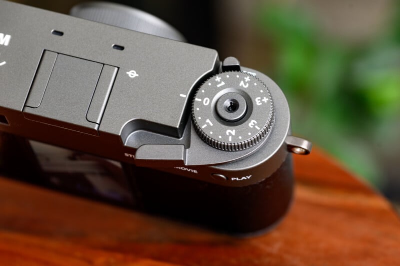Close-up of a camera’s top control dial, showing exposure compensation settings ranging from -3 to +3, with a textured metal finish. The camera rests on a wooden surface with a blurred green background.