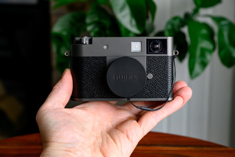 A person holds a Fujifilm camera with a lens cap in their hand, with green leafy plants and a white wall blurred in the background.
