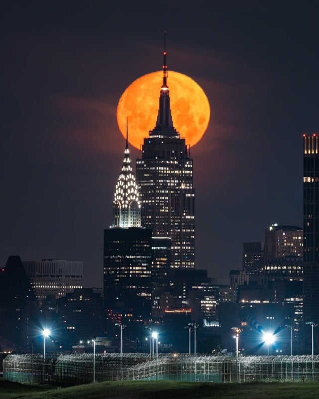 A large orange full moon rises behind New York City’s Empire State Building and Chrysler Building, illuminating the night skyline with glowing city lights in the foreground.
