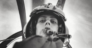 Black and white photo of a pilot wearing a helmet and headset, looking down into the camera from inside a cockpit, with blurred background and cockpit structures visible.